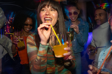 Young, diverse group of friends enjoying a vibrant night out at a busy club, laughing and drinking cocktails while surrounded by colourful neon lights and motion blur creating a dynamic atmosphereの写真素材