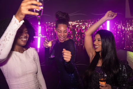 Diverse group of young women dancing and cheering with cocktails in a vibrant nightclub under purple...