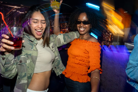Diverse young women are having fun, raising drinks, and dancing together in a lively nightclub...