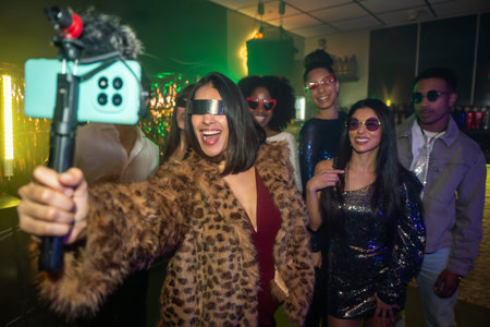 Diverse group of young adults enjoying a night out, capturing fun content with a smartphone on a selfie stick, creating social media buzz at a festive eventの写真素材