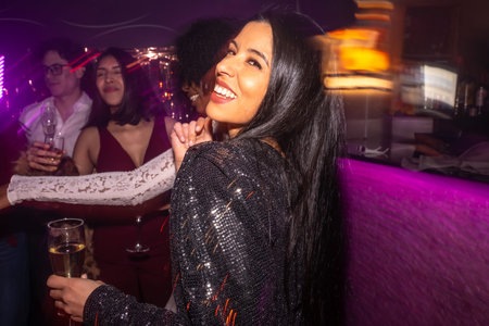 Happy young woman in a sequin dress dancing and smiling with a drink in hand, surrounded by friends...