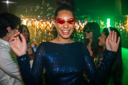 Happy young woman wearing dazzling blue sequin dress and red sunglasses laughing and dancing among friends, celebrating a vibrant party with bubbles, lights, and music in a lively nightclub settingの写真素材