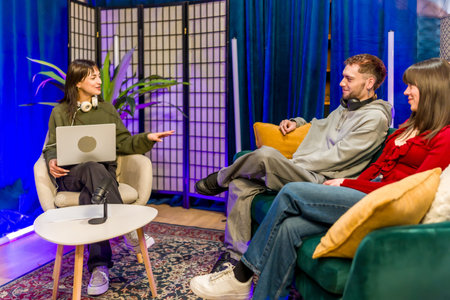 Young diverse creators recording a podcast in a stylish studio, chatting around microphones and laptops, collaborating on digital content for streaming and social media audiencesの写真素材