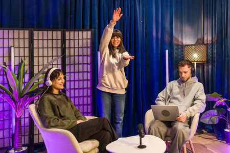 Young adults collaborating in a modern home studio, recording a podcast and creating video content with microphones, laptops and colorful lighting for social media and streaming platformsの写真素材