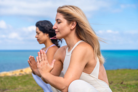 Two women with eyes closed practicing yoga and meditation in prayer pose on the seaside, embracing mindfulness, inner peace and spiritual wellness with ocean horizon and blue skyの写真素材