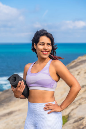 Woman holding a yoga mat and smiling, standing on a rocky cliff overlooking the blue ocean during daytime, enjoying an active and healthy lifestyle while practicing yoga and meditation in natureの写真素材