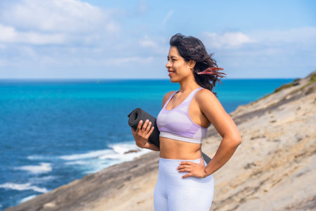 Woman standing on cliff by the ocean, holding a yoga mat and looking to the side, embodying...