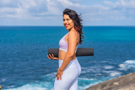 Woman smiling and holding a black yoga mat, standing on a cliff edge with the vast blue ocean and sky creating a serene background, ready for wellness and fitnessの写真素材