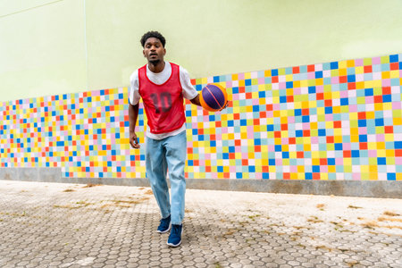 Young black man in casual sportswear playing and dribbling a basketball on a paved court against a...