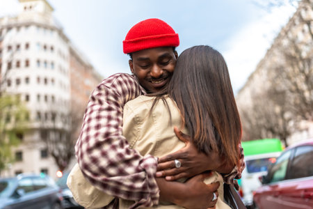 Young man in a red beanie smiles as he embraces his partner on a city street, sharing a warm, candid moment of affection, connection and genuine joy between two peopleの写真素材