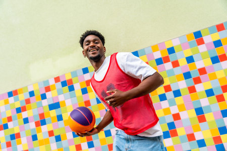 Young black man happily holding a basketball, dressed in a red sports vest and jeans, posing against a vibrant, mosaic tiled background, symbolizing urban youth and active lifestyleの写真素材