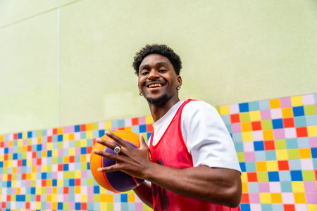 Young man smiling and holding a vibrant basketball in sportswear, standing against a colorful tiled wall outdoors  urban streetball vibe, active lifestyle and joyの写真素材