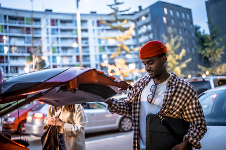 Young man wearing a red beanie smiling while opening a car trunk in an urban parking lot, preparing to load luggage for a trip with another person standing nearbyの写真素材