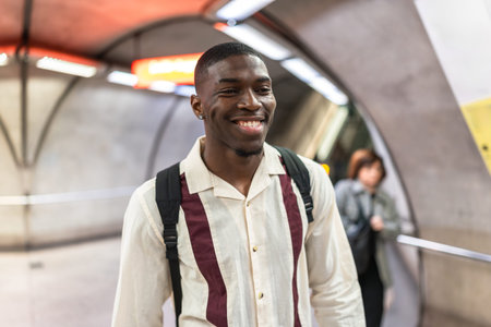 Smiling young african american man with a backpack walking through a modern underground subway station, enjoying his daily commute with a happy and positive expressionの写真素材