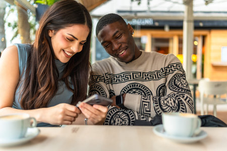 Diverse interracial couple smiling and sharing content on a smartphone at an outdoor cafe, enjoying...