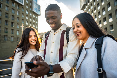 Diverse group of young friends laughing and sharing smartphone content outdoors in a sunny urban setting, enjoying casual connection, social media browsing and togethernessの写真素材