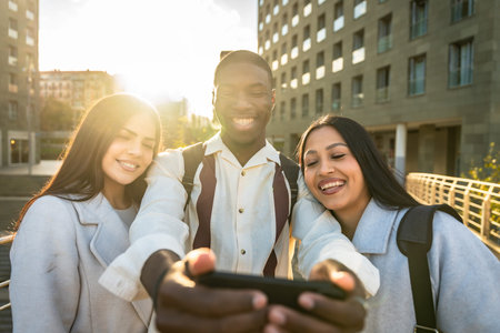 Group of diverse young adults smiling and having fun together while taking a selfie with a smartphone, capturing happy moments in an urban setting with golden hour lightの写真素材