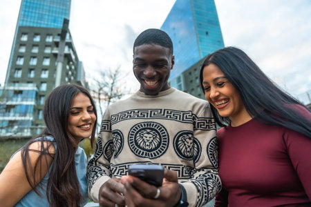 Diverse friends laughing and smiling while gathered on a city street, looking at a smartphone and sharing a joyful, casual moment of connection and social media funの写真素材