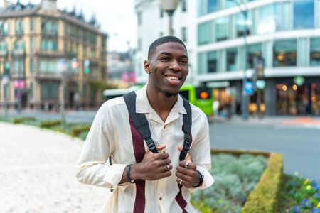 Young black man with a backpack smiling at the camera on a city street, exploring urban architecture...