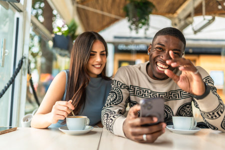 Multiracial couple enjoying a cheerful date, sharing happy moments while viewing media on a mobile phone and drinking coffee in a modern cafeteria settingの写真素材