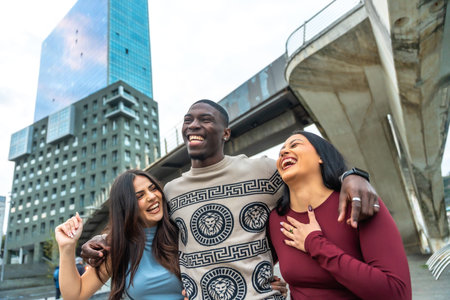 Cheerful diverse group of young adults standing closely together, smiling, laughing, and enjoying a moment of genuine friendship and happiness in a modern city environmentの写真素材