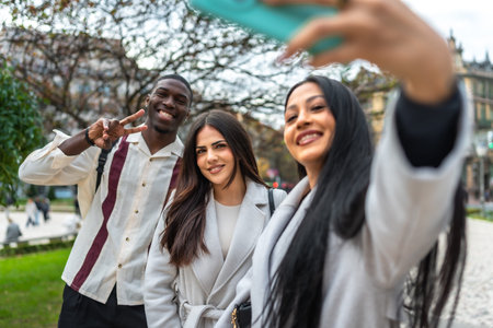 Happy diverse friends smiling and posing for a cheerful selfie with a smartphone outdoors in a city park, capturing a candid moment of youthful fun, connection, and travel memoriesの写真素材