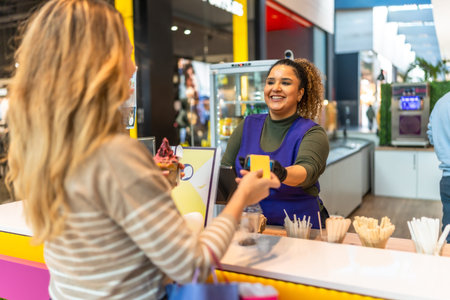 Customer paying with contactless card while smiling cashier completes sweet shop sale at mall counter, friendly service interaction between customer and small business staffの写真素材
