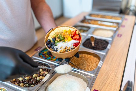 Person in black gloves holding colorful acai bowl with kiwi, blueberries, strawberries and banana while spooning coconut flakes from a counter of topping optionsの写真素材