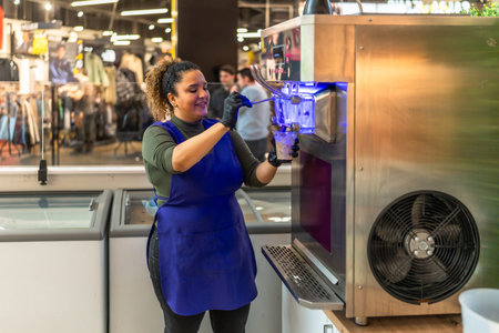 Hispanic woman in blue apron and black gloves smiling while serving soft serve ice cream from a...