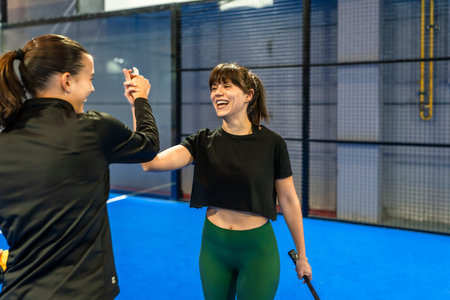 Two young women high fiving on an indoor pickleball court, smiling and celebrating teamwork,...