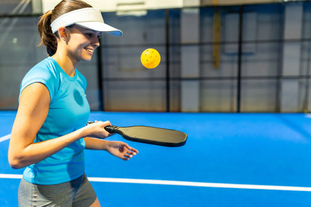 Female athlete smiling while playing pickleball on a blue indoor court, holding a paddle as the ball hovers midair, showing fitness, agility, and energetic recreational playの写真素材