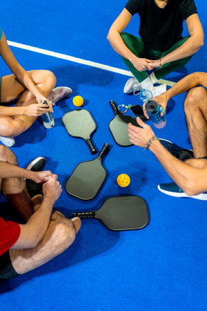 Group of active friends taking a break on a blue pickleball court after playing, holding water...