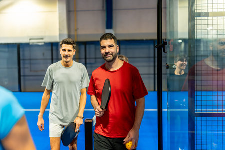 Two smiling men stand on a blue indoor pickleball court holding paddles and a ball, ready to play and enjoy a friendly, active game promoting fitness, fun, and social recreationの写真素材