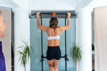 Woman performing a pull up on an exercise bar while a therapist supervises, focusing on...