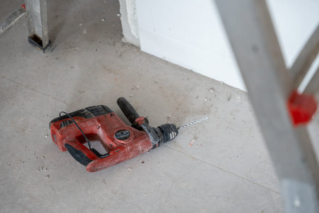 Rotary hammer drill lying on a dusty concrete floor in a room under renovation, indicating ongoing...