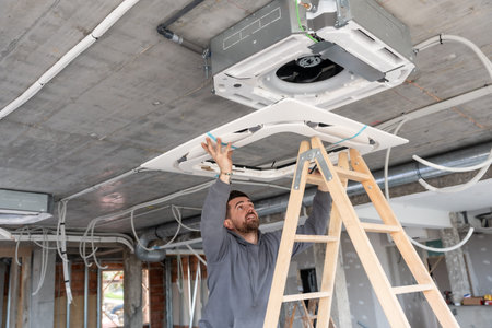 Worker wearing a grey hoodie standing on a wooden ladder, hands reaching up to install a central air...