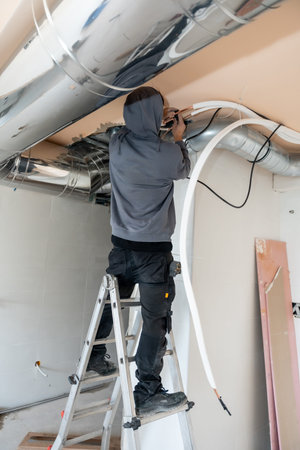 Hvac worker standing on a ladder installing a new air duct and electrical wiring for a climate...