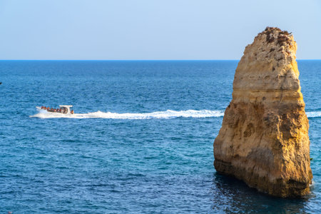 Motorboat full of tourists sailing near a majestic sea stack in the beautiful algarve region of portugal, offering breathtaking views of the coastline and crystal clear watersの写真素材