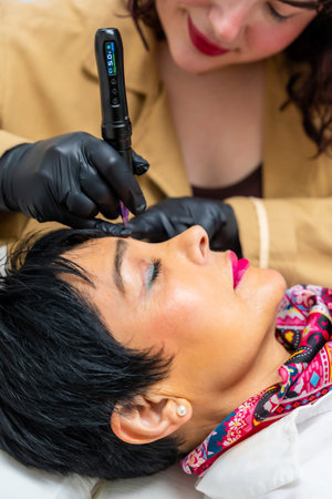 Professional cosmetologist with gloved hands using a specialized pen device, creating microblading...