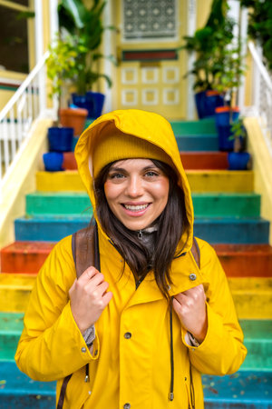 Happy young woman wearing a bright yellow raincoat and beanie smiling at the camera, standing in front of vibrant rainbow painted outdoor steps in a colorful urban neighborhood