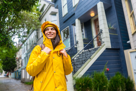 Woman in a bright yellow raincoat and hood, smiling while exploring a street with classic colorful victorian style houses during an overcast day in san francisco