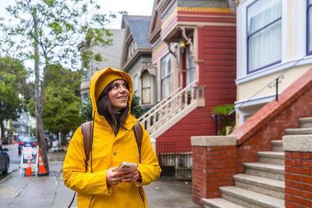 Woman in yellow raincoat and beanie walking a san francisco street of colorful victorian and edwardian homes, smiling and exploring the historic neighborhood with phone in hand