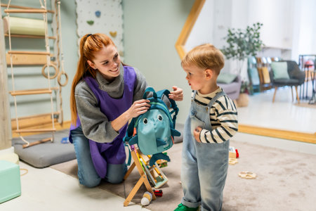 Smiling preschool teacher assisting a young boy with his shark themed backpack, preparing for the day or departure in a bright and playful kindergarten classroom