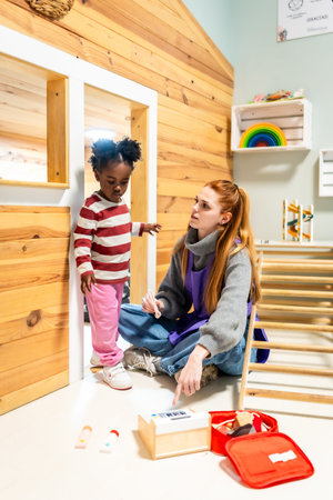 Young woman teacher sitting on the floor interacting with a little black girl toddler playing with a toy medical kit inside a wooden playhouse at a daycare