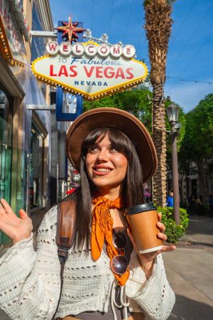 Young woman smiling and holding a coffee cup, standing on a sunny street with a famous welcome sign and palm trees, exploring the vibrant city of las vegas