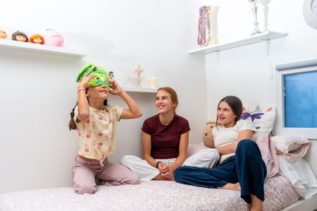 Young girls wearing pajamas enjoying a fun sleepover, one girl trying on a green novelty sleep mask while her two friends smile and laugh, bonding during childhood moments indoors