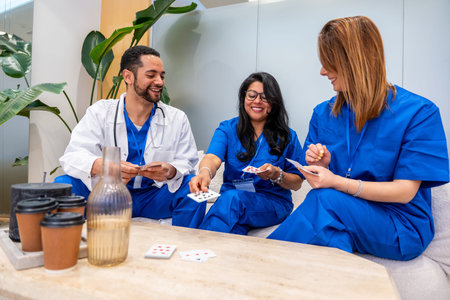 Group of smiling doctors and nurses in blue scrubs and white lab coat engaging in a casual card game, sharing moments of joy and stress relief during a work break in a modern hospital common room