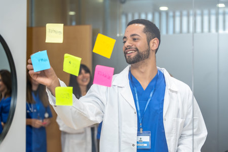 Doctor in medical uniform and lab coat brainstorming and organizing ideas on a glass wall with colorful sticky notes, colleagues smiling in background, focusing on healthcare concepts