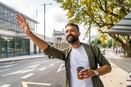 Bearded man in smart casual attire holds a coffee cup and raises his hand on a sunny city sidewalk to hail a taxi, smiling as he waits for his urban commute