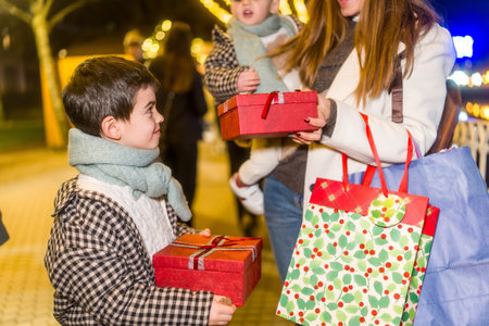 Mother holding a baby and presenting a carefully wrapped red gift to her happy child while standing outdoors with shopping bags during the holiday season